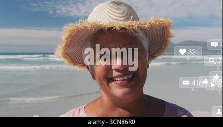 Femme senior souriante portant chapeau de soleil de paille et haut floral rose à la plage, icônes de réaction flottantes Banque D'Images