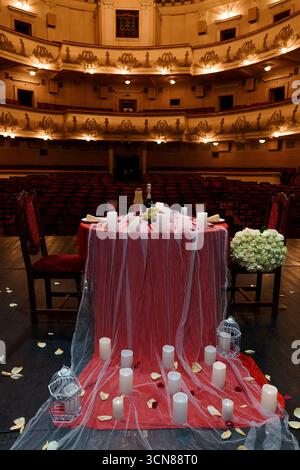 Un dîner romantique aux chandelles mis en place pour deux dans un théâtre vintage vide. la table drapée de rouge avec des roses, du champagne et un éclairage doux crée un i. Banque D'Images