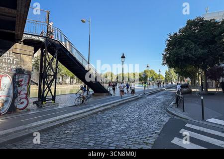 Coureurs, piétons et joggeurs profitent d'une journée beuatifiante le long du canal de l'Ourcq dans le 19ème arrondissement de Paris le 7 septembre 2025. Un large canal urbain ou une rivière coule entre deux promenades bordées d'arbres dans un cadre urbain. A droite, une large passerelle en pierre est bordée par une balustrade et des piétons ; à gauche, un bas bord de pierre mène à l'eau. Au loin, des arbres et un groupe de bâtiments de faible hauteur et une grande structure blanche moderne s'élèvent contre un ciel bleu clair. La scène semble calme et ensoleillée, avec la lumière qui se reflète sur l'eau. (Photo de Samuel Rigelhaupt/Sipa USA) Banque D'Images