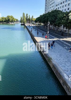 Coureurs, piétons et joggeurs profitent d'une journée beuatifiante le long du canal de l'Ourcq dans le 19ème arrondissement de Paris le 7 septembre 2025. Un large canal urbain ou une rivière coule entre deux promenades bordées d'arbres dans un cadre urbain. A droite, une large passerelle en pierre est bordée par une balustrade et des piétons ; à gauche, un bas bord de pierre mène à l'eau. Au loin, des arbres et un groupe de bâtiments de faible hauteur et une grande structure blanche moderne s'élèvent contre un ciel bleu clair. La scène semble calme et ensoleillée, avec la lumière qui se reflète sur l'eau. (Photo de Samuel Rigelhaupt/Sipa USA) Banque D'Images