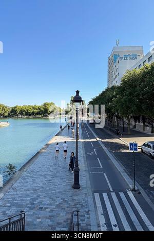 Coureurs, piétons et joggeurs profitent d'une journée beuatifiante le long du canal de l'Ourcq dans le 19ème arrondissement de Paris le 7 septembre 2025. Un large canal urbain ou une rivière coule entre deux promenades bordées d'arbres dans un cadre urbain. A droite, une large passerelle en pierre est bordée par une balustrade et des piétons ; à gauche, un bas bord de pierre mène à l'eau. Au loin, des arbres et un groupe de bâtiments de faible hauteur et une grande structure blanche moderne s'élèvent contre un ciel bleu clair. La scène semble calme et ensoleillée, avec la lumière qui se reflète sur l'eau. (Photo de Samuel Rigelhaupt/Sipa USA) Banque D'Images