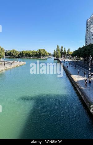 Coureurs, piétons et joggeurs profitent d'une journée beuatifiante le long du canal de l'Ourcq dans le 19ème arrondissement de Paris le 7 septembre 2025. Un large canal urbain ou une rivière coule entre deux promenades bordées d'arbres dans un cadre urbain. A droite, une large passerelle en pierre est bordée par une balustrade et des piétons ; à gauche, un bas bord de pierre mène à l'eau. Au loin, des arbres et un groupe de bâtiments de faible hauteur et une grande structure blanche moderne s'élèvent contre un ciel bleu clair. La scène semble calme et ensoleillée, avec la lumière qui se reflète sur l'eau. (Photo de Samuel Rigelhaupt/Sipa USA) Banque D'Images
