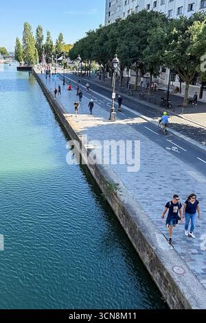 Coureurs, piétons et joggeurs profitent d'une journée beuatifiante le long du canal de l'Ourcq dans le 19ème arrondissement de Paris le 7 septembre 2025. Un large canal urbain ou une rivière coule entre deux promenades bordées d'arbres dans un cadre urbain. A droite, une large passerelle en pierre est bordée par une balustrade et des piétons ; à gauche, un bas bord de pierre mène à l'eau. Au loin, des arbres et un groupe de bâtiments de faible hauteur et une grande structure blanche moderne s'élèvent contre un ciel bleu clair. La scène semble calme et ensoleillée, avec la lumière qui se reflète sur l'eau. (Photo de Samuel Rigelhaupt/Sipa USA) Banque D'Images