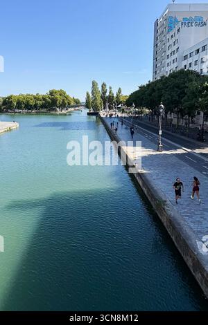 Coureurs, piétons et joggeurs profitent d'une journée beuatifiante le long du canal de l'Ourcq dans le 19ème arrondissement de Paris le 7 septembre 2025. Un large canal urbain ou une rivière coule entre deux promenades bordées d'arbres dans un cadre urbain. A droite, une large passerelle en pierre est bordée par une balustrade et des piétons ; à gauche, un bas bord de pierre mène à l'eau. Au loin, des arbres et un groupe de bâtiments de faible hauteur et une grande structure blanche moderne s'élèvent contre un ciel bleu clair. La scène semble calme et ensoleillée, avec la lumière qui se reflète sur l'eau. (Photo de Samuel Rigelhaupt/Sipa USA) Banque D'Images