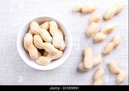 Cacahuètes grillées en coquille dans un bol blanc sur lin. Noix entières croquantes, fruits d'Arachis hypogaea enfermés dans sa coquille naturelle. Banque D'Images