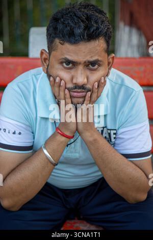 Un jeune Indien est assis sur un banc de parc, posant son menton sur ses mains avec une expression sérieuse. La lumière du jour brillante met en valeur son réfléchi et reflec Banque D'Images