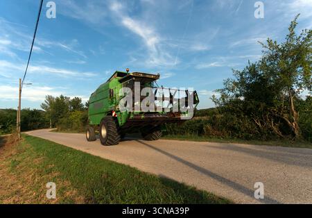 Une grande moissonneuse-batteuse verte emprunte une route de campagne étroite et sinueuse, laissant un petit sillage de poussière sur son passage. Banque D'Images