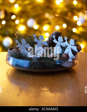 Couronne de l'Avent simple faite à la main avec quatre anges en métal blanc, branches de pin vert et des noix dans un bol en verre sur une table en bois avec arbre de Noël flou Banque D'Images