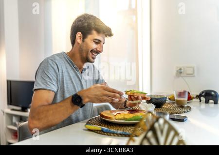 Jeune homme souriant tout en profitant d'un petit déjeuner nutritif de pain grillé à l'avocat dans une cuisine élégante et moderne, embrassant un mode de vie sain Banque D'Images