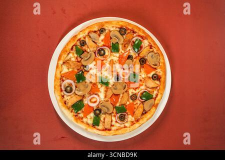 Un cliché studio en grand angle d'une délicieuse pizza au poulet et aux champignons fraîchement cuite avec des poivrons et des oignons sur une assiette avec un fond rouge. Banque D'Images