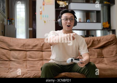 Un jeune homme est assis sur un canapé marron dans un salon, portant des écouteurs et des lunettes, tenant une manette de jeu et pointant vers un écran avec un Banque D'Images