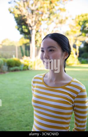 Femme asiatique debout dans le jardin ensoleillé de l'arrière-cour portant une chemise rayée à manches longues, espace copie Banque D'Images