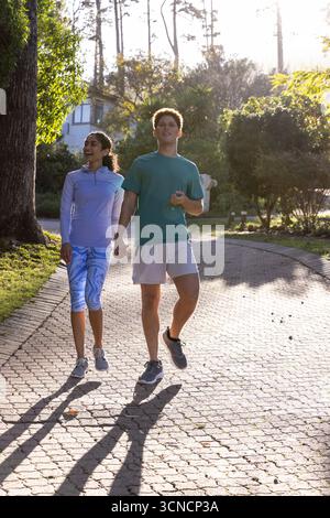 Couple diversifié marchant tenant les mains sur le chemin de brique dans le parc ensoleillé portant des chaussures Banque D'Images