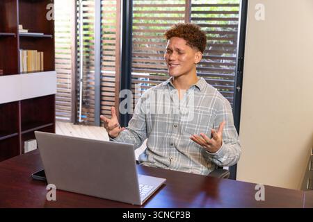 Travailleur de bureau masculin gestuel, parlant pendant la réunion en ligne dans l'étude à domicile avec ordinateur portable, bibliothèque Banque D'Images
