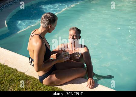 Couple senior en maillots de bain et bikini se relaxant à la terrasse de la piscine arrière tenant des lunettes Banque D'Images