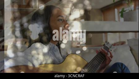 Musicien de guitare acoustique strumming portant une tenue en denim sur le canapé dans le salon, avec des lumières bokeh Banque D'Images