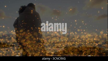 Couple serrant debout enveloppé dans une couverture sur la plage au coucher du soleil, avec des vagues de l'océan et bokeh doré Banque D'Images