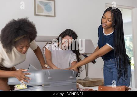 Divers amis féminins emballant des articles de vêtements dans la valise en argent sur le lit dans la chambre à coucher Banque D'Images
