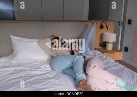 Couple féminin diversifié dans des vêtements de détente pastel couché sur le lit avec couette blanche dans la chambre Banque D'Images