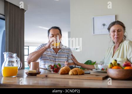 Couple senior diversifié partageant le petit déjeuner, buvant du jus d'orange et mangeant des croissants Banque D'Images