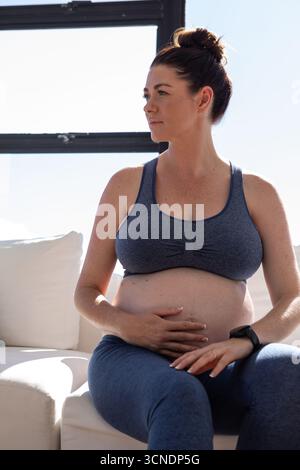 Femme enceinte blanche berçant la bosse assise sur le canapé portant des vêtements de sport bleus avec tracker près de la fenêtre Banque D'Images