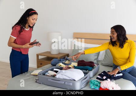 Maman diversifiée et fille adolescente organisant des vêtements dans une valise grise sur le lit avec smartphone Banque D'Images