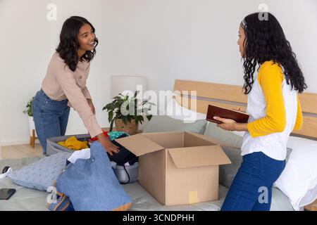 Maman diversifiée et fille adolescente debout près du lit emballant des vêtements de la valise ouverte dans la boîte Banque D'Images