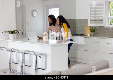 Maman diversifiée et fille adolescente debout à l'îlot de cuisine pointant vers l'ordinateur portable et tenant des papiers Banque D'Images