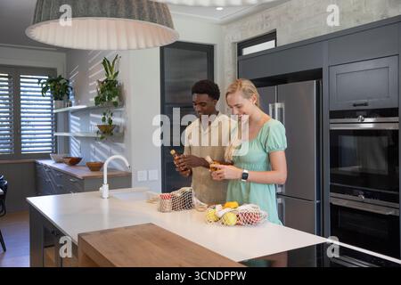 Couple diversifié déballer l'épicerie et tenir des bouteilles en verre à l'îlot de cuisine avec des sacs en filet Banque D'Images