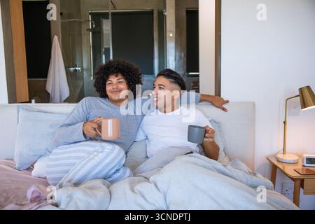 Hommes divers portant un pyjama, appuyés contre la tête de lit, sirotant du café dans des tasses dans la chambre principale Banque D'Images