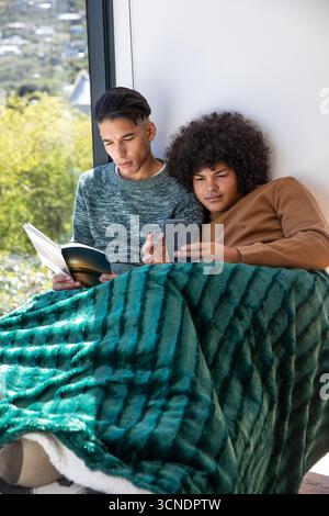 Divers amis masculins assis sur le siège de fenêtre lisant livre de poche et vérifiant smartphone à la maison Banque D'Images