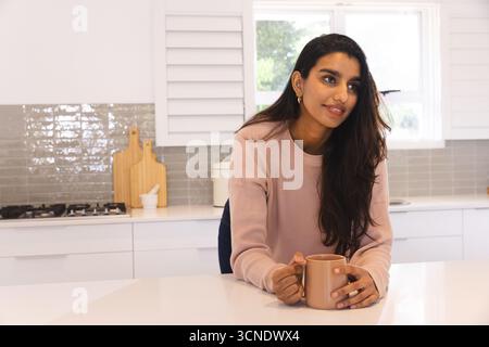 Femme indienne dans ses années 30 s'appuyant sur le comptoir de quartz dans la cuisine tenant une tasse à café, espace de copie Banque D'Images