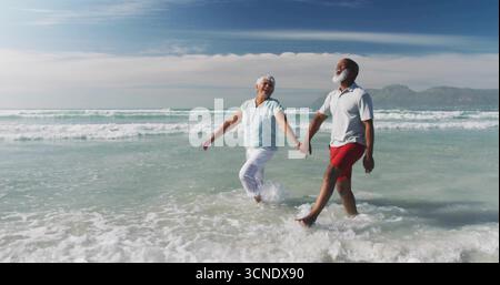 Promenez-vous les retraités en vêtements de plage à travers le surf sur la rive sablonneuse avec les vagues de l'océan et les collines lointaines Banque D'Images