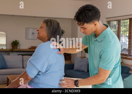 Femme indienne senior recevant le massage du dos du soignant masculin dans le salon avec canapé, plantes Banque D'Images
