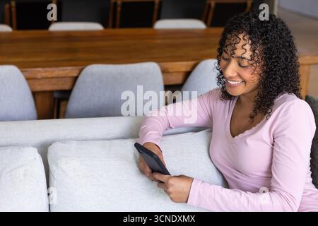 Femme afro-américaine assise sur le canapé défilant smartphone et souriant à l'écran, espace de copie Banque D'Images
