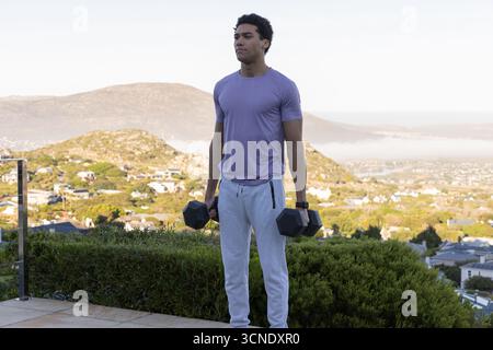 Homme afro-américain portant un tracker saisissant deux haltères sur le patio par la balustrade face à la vallée Banque D'Images