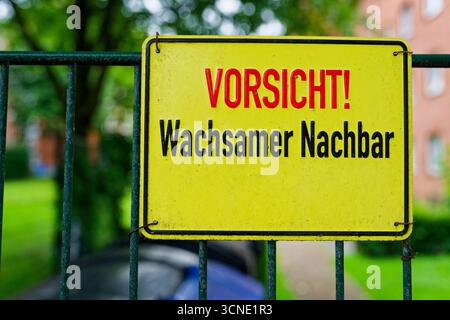 Une clôture métallique basse fait face à une rangée de poubelles, tandis qu'un panneau d'avertissement jaune en allemand est accroché à proximité. Un bâtiment plat flanque la scène, le sol humide, l'ai Banque D'Images