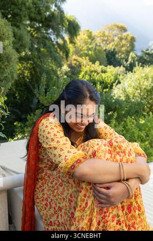 Femme indienne portant kurta jaune, écharpe rouge assise sur le rebord du balcon serrant les genoux, souriant Banque D'Images