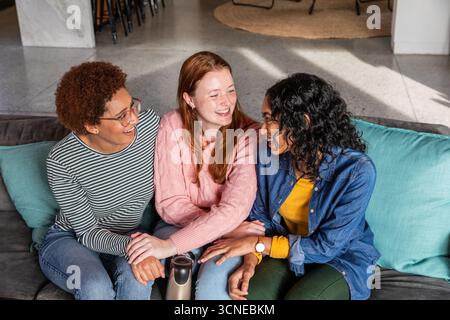 Diverses amies féminines riant et se penchant sur le canapé dans le salon avec flacon thermos en acier Banque D'Images
