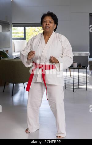 Femme indienne senior pratiquant les arts martiaux dans le salon portant GI blanc et ceinture rouge Banque D'Images