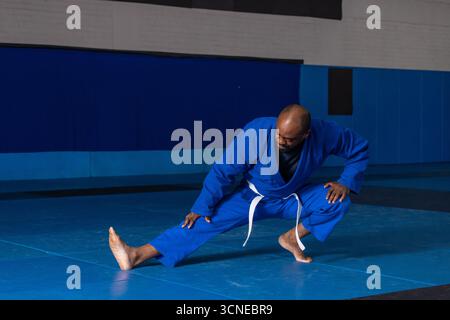 Homme afro-américain en GI bleu, ceinture blanche effectuant une fente latérale sur des tapis avant des panneaux rembourrés Banque D'Images