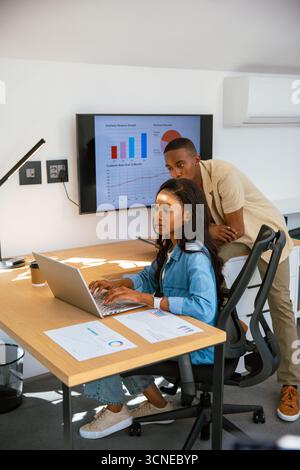 Collègues afro-américains examinant les graphiques trimestriels sur ordinateur portable et moniteur au bureau Banque D'Images