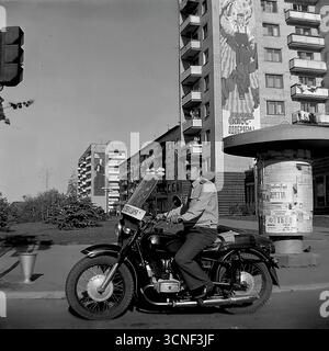 Un milicien soviétique, un officier de la GAI (State automobile Inspectorate), est assis sur sa moto de patrouille alors qu'il est en service à un carrefour de la ville de Sloviansk, RSS d'Ukraine, lors de la célébration de la fête de la ville le 6 septembre 1985. Le fond urbain authentique comprend des immeubles à panneaux typiques de l'ère soviétique décorés d'énormes peintures murales de propagande, dont une avec le slogan « nous approuvons la politique du PCUS ! », créant un instantané puissant et atmosphérique de l'application de la loi et de l'idéologie à la fin de l'URSS Banque D'Images