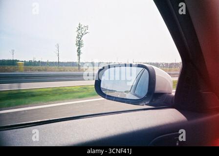 Photo d'archive analogique d'un paysage de terres agricoles vu d'une voiture en mouvement. Le rétroviseur latéral au premier plan reflète la route derrière, ajoutant de la profondeur. Banque D'Images