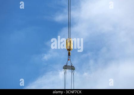 Le mécanisme de levage jaune de Crane, suspendu en l'air par deux câbles métalliques, supporte une dalle rectangulaire grise placée contre un ciel bleu vif partiellement nuageux. Banque D'Images