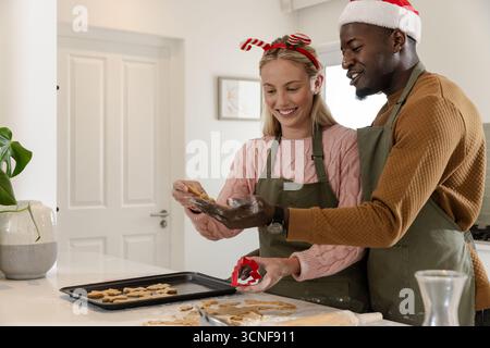 Couple diversifié faisant des biscuits étoilés sur le comptoir de cuisine dans des tabliers avec des couteaux et un plateau de cuisson Banque D'Images