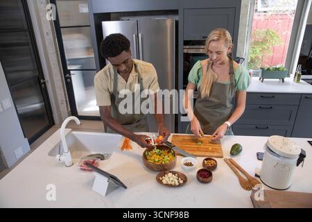 Couple diversifié portant des tabliers épluchant des carottes sur un bol, tranchant des concombres sur l'île dans la cuisine Banque D'Images