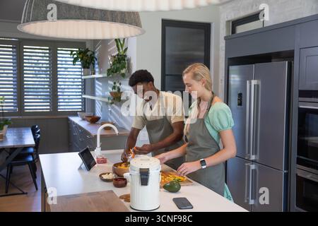 Couple diversifié dans des tabliers hachant des légumes sur une planche à découper à l'îlot de cuisine avec tablette Banque D'Images