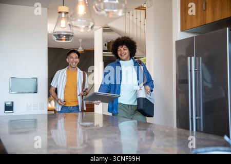 Divers amis masculins marchant dans la cuisine par île de pierre portant smartphone et ordinateur portable Banque D'Images