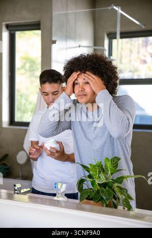 Divers amis masculins debout à l'évier de salle de bain appliquant la crème pour le visage tout en ouvrant le pot hydratant Banque D'Images